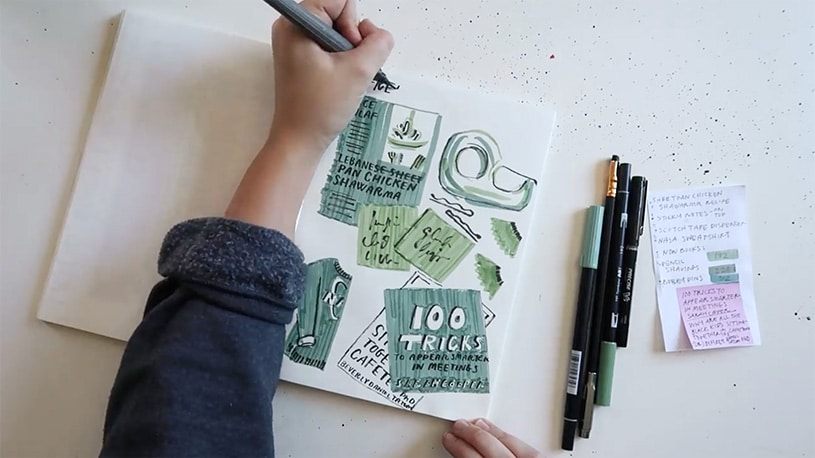 A hand drawing in a sketchbook filled with green illustrations and notes, with pens and sticky notes nearby.